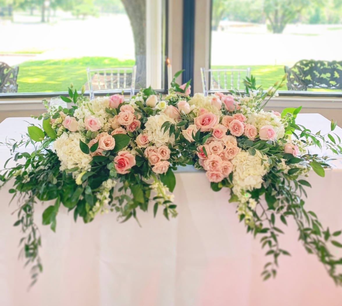a bridal sweet heart table with a large, overspilling flower arrangement on the front edge of the table. The flowers are roses, hydrangeas and carnations in pink and creme with Italian ruscus, ivy and salal greeneries.